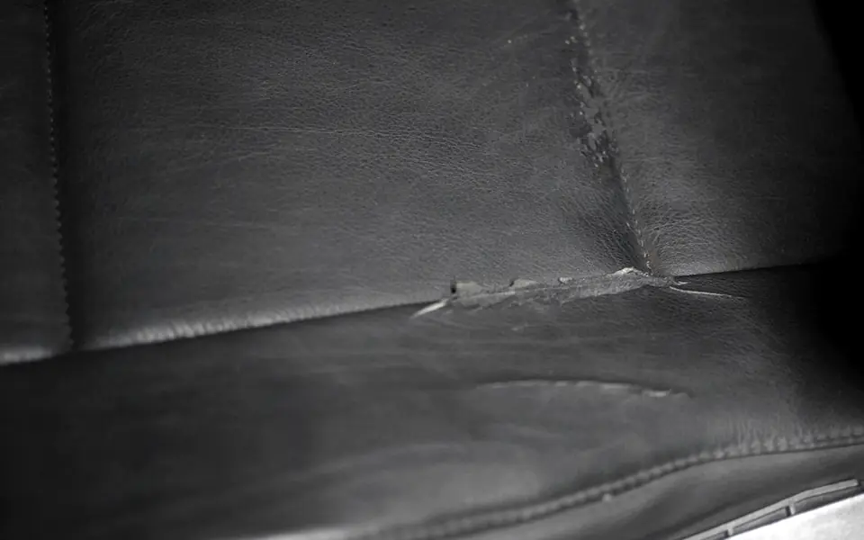 A worn black leather seat where the hide has split open along the seam between the backrest and the cushion. The edges of the tear have curled and gone grey, and the grain either side of it has lost its sheen.