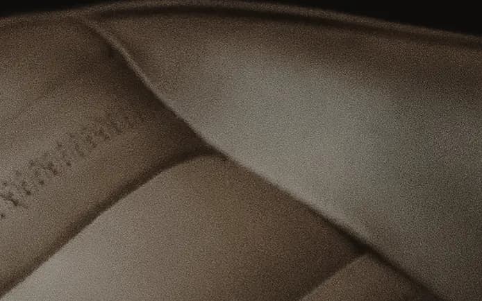 A tan leather seat cushion in soft light, divided into rounded flutes. A line of small punched holes runs along the valley between two flutes, the perforation that lets a hide breathe.