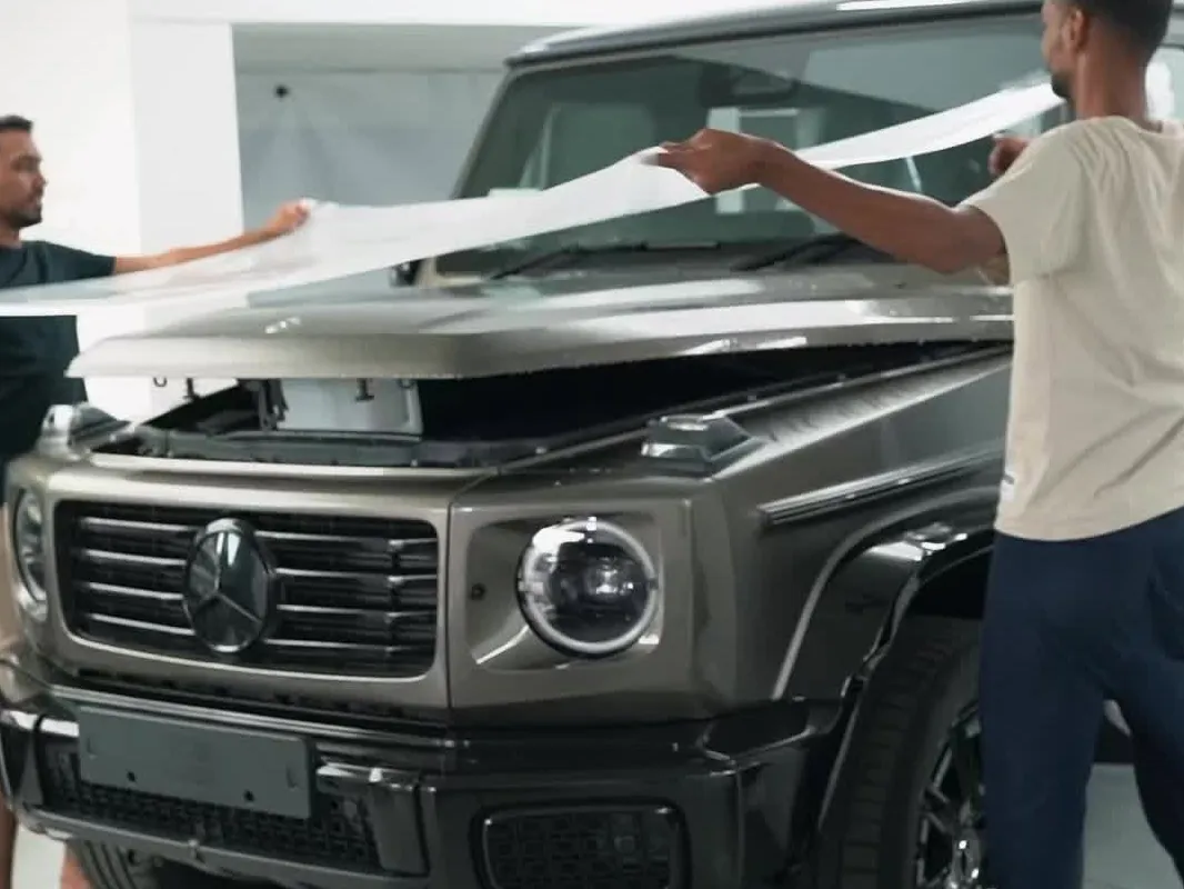 Two technicians laying a sheet of paint protection film across the bonnet of a silver Mercedes G-Class